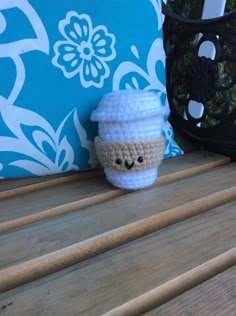 a crocheted coffee cup sitting on top of a wooden bench next to a pillow