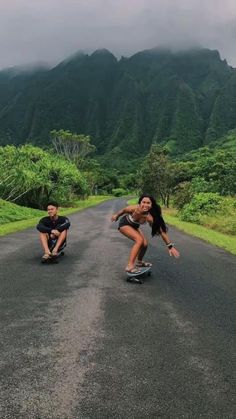 two people riding skateboards down a road in the middle of mountains and grass on both sides