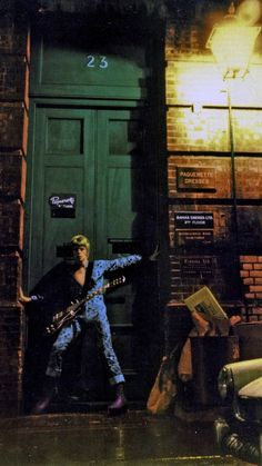 a man with a guitar standing in front of a green door on a brick building