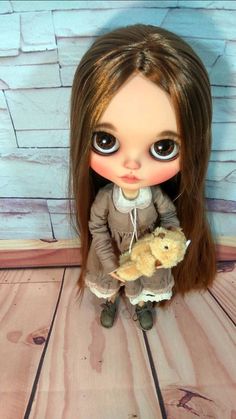 a doll with long brown hair and big eyes holding a teddy bear in front of a brick wall