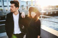 a man and woman standing next to each other on a bridge near the water with buildings in the background