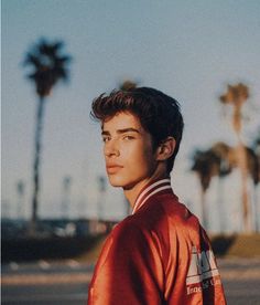 a young man standing in front of palm trees wearing a red jacket and white collared shirt