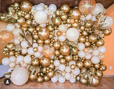 gold and white balloons are arranged on the floor in front of a wall that is decorated with greenery