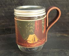 a mason jar with a handle is sitting on a table next to a mug that has a camp scene painted on it