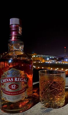 a bottle of chivas regal next to a glass on a table with lights in the background