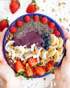 two hands holding a blue bowl filled with fruit and granola on top of it