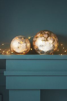 two shiny balls sitting on top of a mantle with christmas lights around them in front of a blue wall