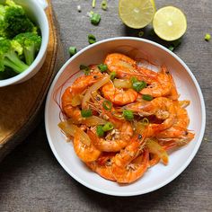 a white bowl filled with cooked shrimp next to broccoli and lemon wedges