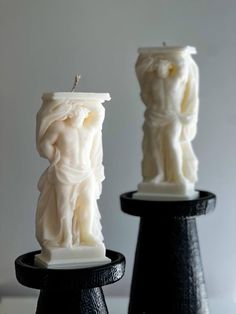 two white candles sitting next to each other on top of black pedestals with one candle in the shape of a statue