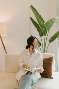 a woman is sitting on a couch reading a book and smiling at the camera, with a plant in the background