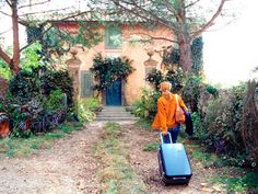 a woman with a suitcase is walking towards a house in the country side garden area