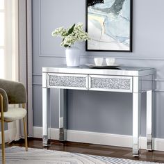 a white console table with two drawers and a vase on the top, in front of a blue wall