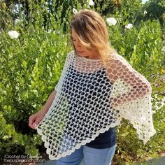 a woman wearing a white crocheted shawl in front of some bushes and flowers