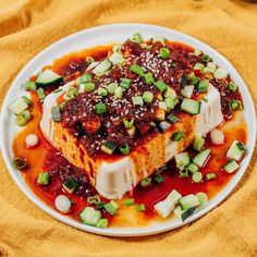 a white plate topped with tofu covered in sauce and green onions on top of a yellow cloth