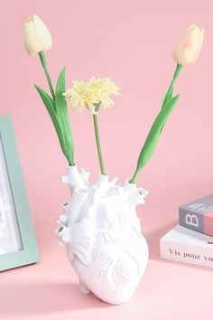 three white flowers in a vase next to books and a framed photograph on a pink background