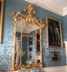 an ornate gold framed mirror in the corner of a room with blue wallpaper and paintings