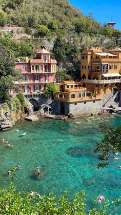 people are swimming in the clear blue water near some buildings and trees on top of a hill