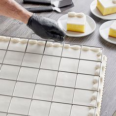 a person in black gloves is decorating a cake with white frosting and square squares