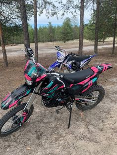 a pink and blue dirt bike parked on top of a dirt field next to trees