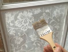 a person holding a paint brush in front of a window with white lace on it