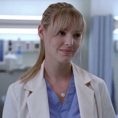 a woman with blonde hair wearing a white coat and smiling at the camera in a hospital room