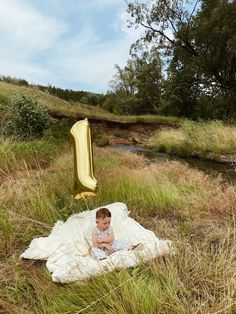 a baby is sitting on a blanket in the grass near a stream with a gold balloon