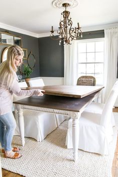 a woman standing in front of a dining room table with white chairs and a chandelier