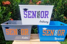 three bins with different colors and logos on them sitting on a bench in front of some bushes