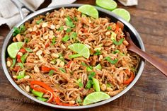 a pan filled with noodles and vegetables on top of a wooden table next to lime wedges