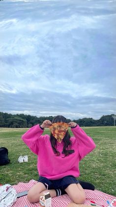 a woman sitting on top of a picnic blanket holding a slice of pizza over her face