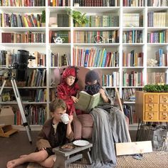 two children sitting on a chair in front of bookshelves with one reading and the other drinking from a cup