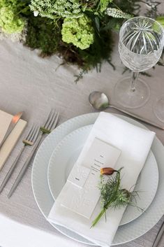 the table is set with white plates and silverware, greenery, and wine glasses