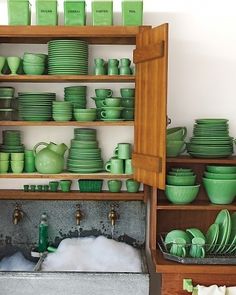 green dishes are stacked on wooden shelves in front of white walls and open cupboards