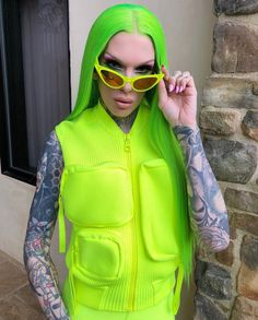 a woman with green hair and tattoos wearing neon yellow clothing, holding her sunglasses up to her face