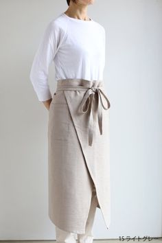 a woman standing in front of a wall wearing a white shirt and beige skirt with a tie around the waist