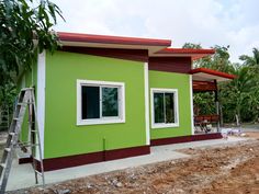 a green and red house being built on the side of a dirt road with trees in the background