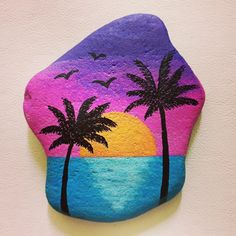 a painted rock with two palm trees and the sun setting in the sky behind it