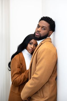 a man and woman standing next to each other in front of a white wall with their arms around each other