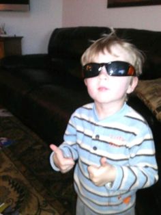 a young boy wearing sunglasses standing in front of a couch with his hand up to the camera