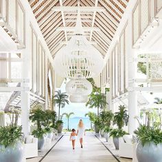 a woman walking down a long hallway between two large white vases filled with plants