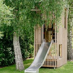 a wooden structure with a slide in front of it and trees surrounding it on the grass