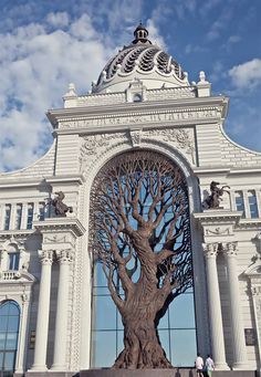 a large white building with a giant tree in the center and people walking around it