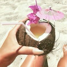 a person holding a coconut drink in their hand on the beach with an umbrella over it