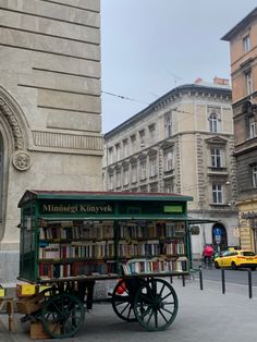 an old fashioned cart with books on it in front of a stone building and people walking by