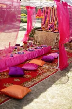 a table set up for a party with pink linens and pillows on the ground