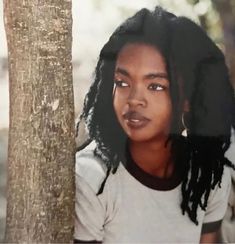 a woman with dreadlocks standing next to a tree in front of a blurry background