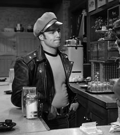 a man in a leather jacket standing next to a woman at a counter with glasses