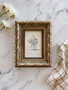 an ornate gold frame holds a drawing of a vase with flowers in it on a marble surface