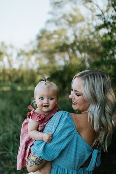 a woman holding a baby in her arms and smiling at the camera with trees in the background