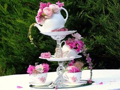 three tiered cake stand with pink flowers and teacups on the top, surrounded by greenery